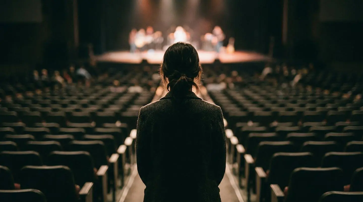Silhouette d'une personne vue de dos face à un auditorium aux rangées de sièges floutées, légère contre-jour soulignant la posture d'attente avant intervention